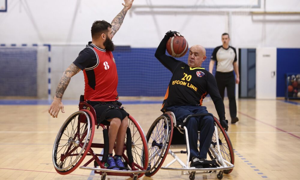 Fotogracija prikazuje dva igrača košarke u kolicima. Jedan drži podignutu ruku, a drugi vodi loptu.
