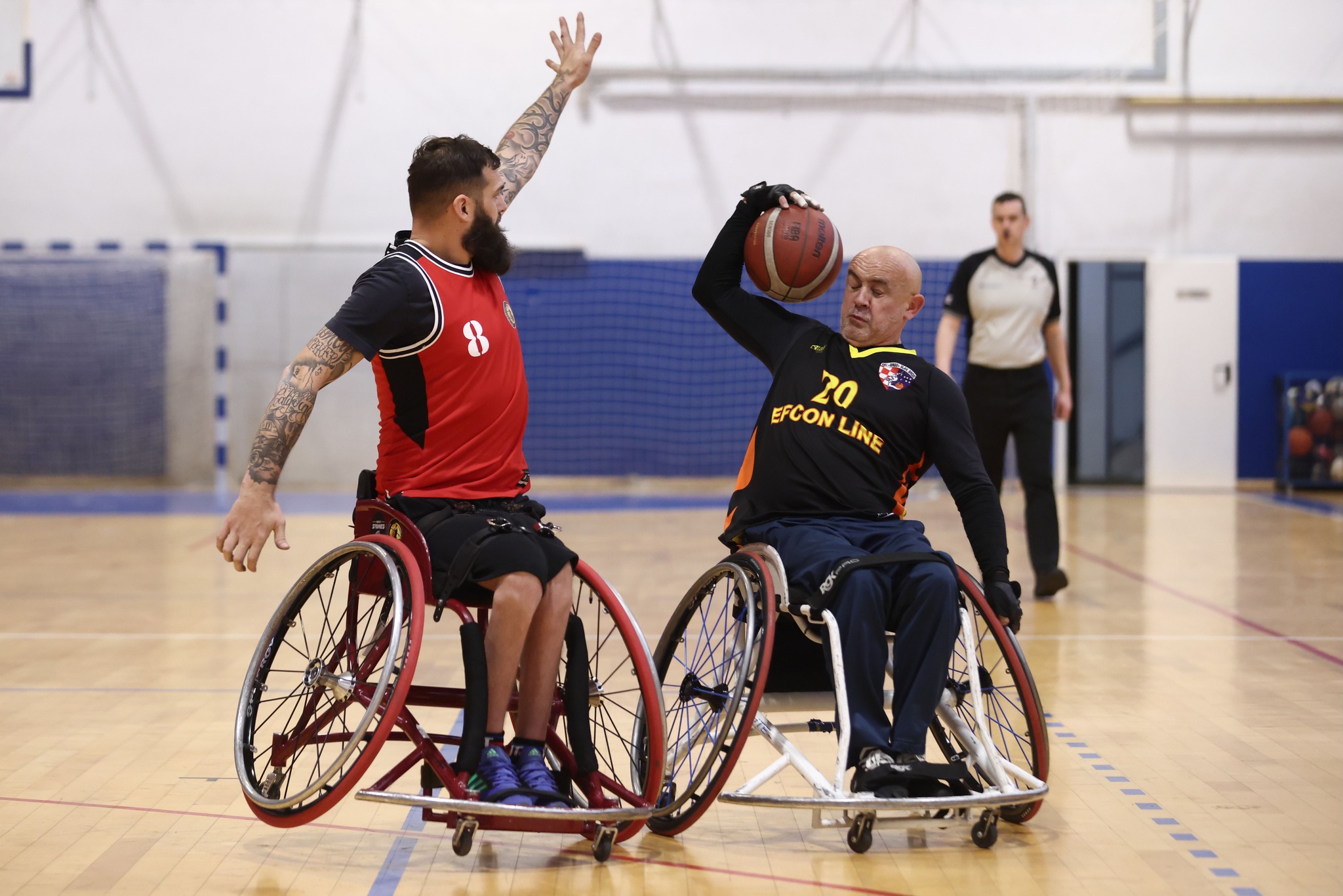Fotogracija prikazuje dva igrača košarke u kolicima. Jedan drži podignutu ruku, a drugi vodi loptu.