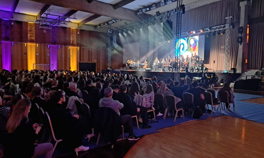 Puna koncertna dvorana s publikom koja sjedi i gleda prema velikoj pozornici na kojoj nastupa glazbeni ansambl s orkestrom i pjevačima. Pozornica je osvijetljena snažnim reflektorima i velikim LED ekranom u pozadini, dok u publici vlada svečana i pažljiva atmosfera.