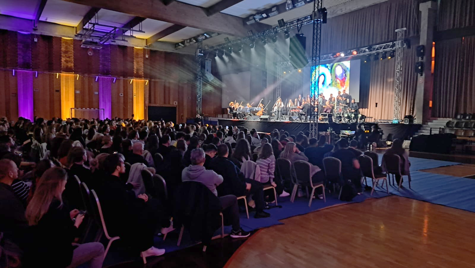 Puna koncertna dvorana s publikom koja sjedi i gleda prema velikoj pozornici na kojoj nastupa glazbeni ansambl s orkestrom i pjevačima. Pozornica je osvijetljena snažnim reflektorima i velikim LED ekranom u pozadini, dok u publici vlada svečana i pažljiva atmosfera.
