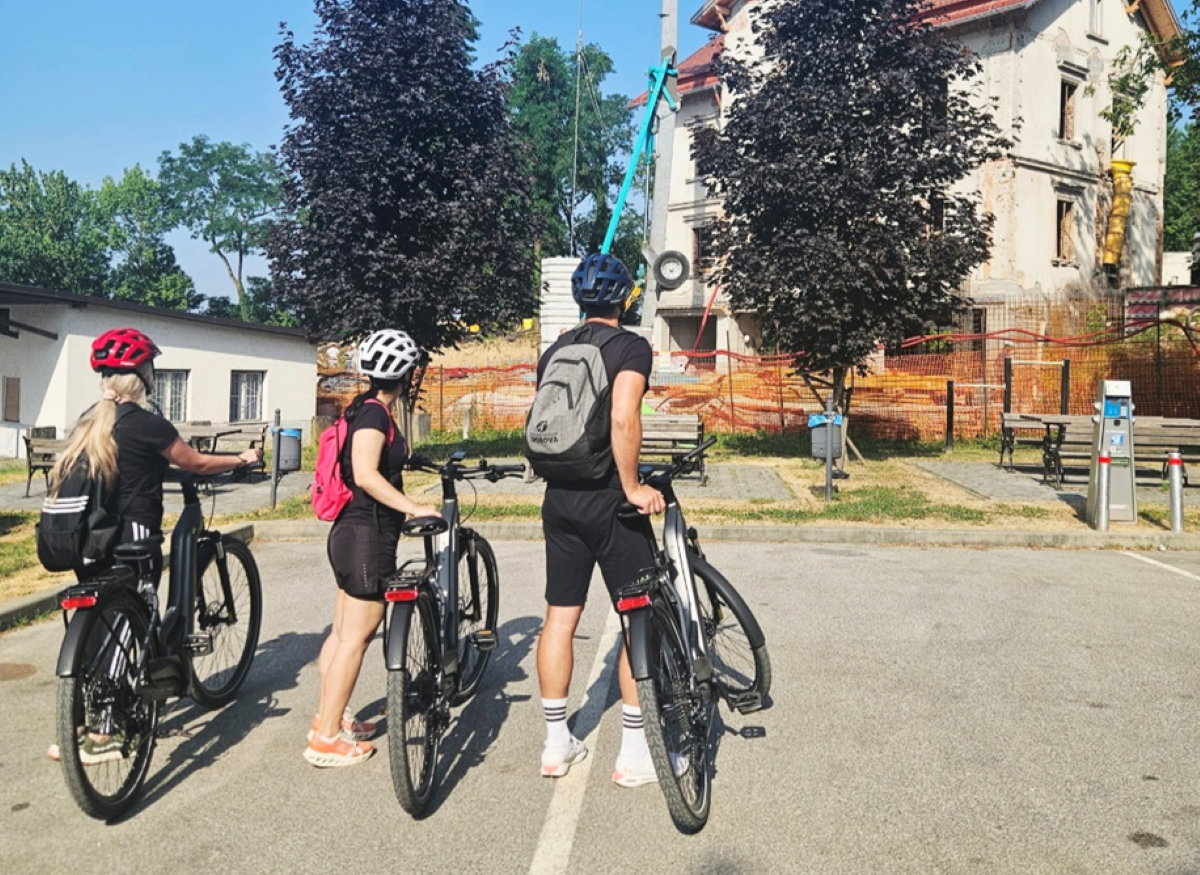 Na fotografiji su tri osobe s biciklima, zaustavljene na popločanom prostoru. Svi nose kacige i ruksake, a pogled im je usmjeren prema zgradi koja je očito u fazi obnove ili izgradnje. Zgrada je ograđena narančastom zaštitnom mrežom, a oko nje se nalaze građevinski elementi poput dizalice i cijevi za odvoz šute. Na fasadi su vidljivi tragovi građevinskih radova – otvoreni zidovi, skele i drugi znakovi konstrukcije. Okruženje je sunčano, s drvećem i klupama u blizini, što sugerira da se scena odvija u urbanom parku ili javnom prostoru. Cijeli prizor odiše znatiželjom i promišljanjem – kao da biciklisti zastaju kako bi promotrili promjene u svom gradu, možda tijekom rekreativne vožnje ili vođenog obilaska.
