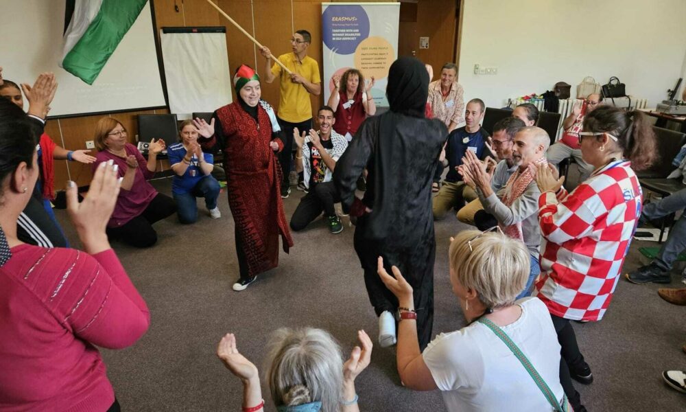 Fotografija prikazuje živopisan trenutak zajedništva i kulturne razmjene u zatvorenom prostoru, vjerojatno tijekom društvenog ili edukativnog događanja. U središtu su dvije osobe u tradicionalnoj nošnji koje plešu, dok ih okupljeni sudionici okružuju pljeskom i vedrim izrazima lica. Atmosfera je dinamična i inkluzivna. Jedna osoba drži palestinsku zastavu, a druga pokazuje štapom prema natpisu na pozadini koji glasi: ERASMUS+ ZAJEDNO S RAZLIČITIMA I UNUTAR RAZLIČITOSTI ZAGOVARAMO SEBE! VODI SVOJE VRŠNJAKE! SUDJELUJ U SVOJOJ ZAJEDNICI! NEKA SE TVOJ GLAS ČUJE! BUDI VIDLJIV! Ova scena snažno prenosi poruku o važnosti samozastupanja, sudjelovanja i vidljivosti unutar raznolikih zajednica, u duhu programa Erasmus+ koji promiče inkluziju, kulturnu razmjenu i aktivno građanstvo.