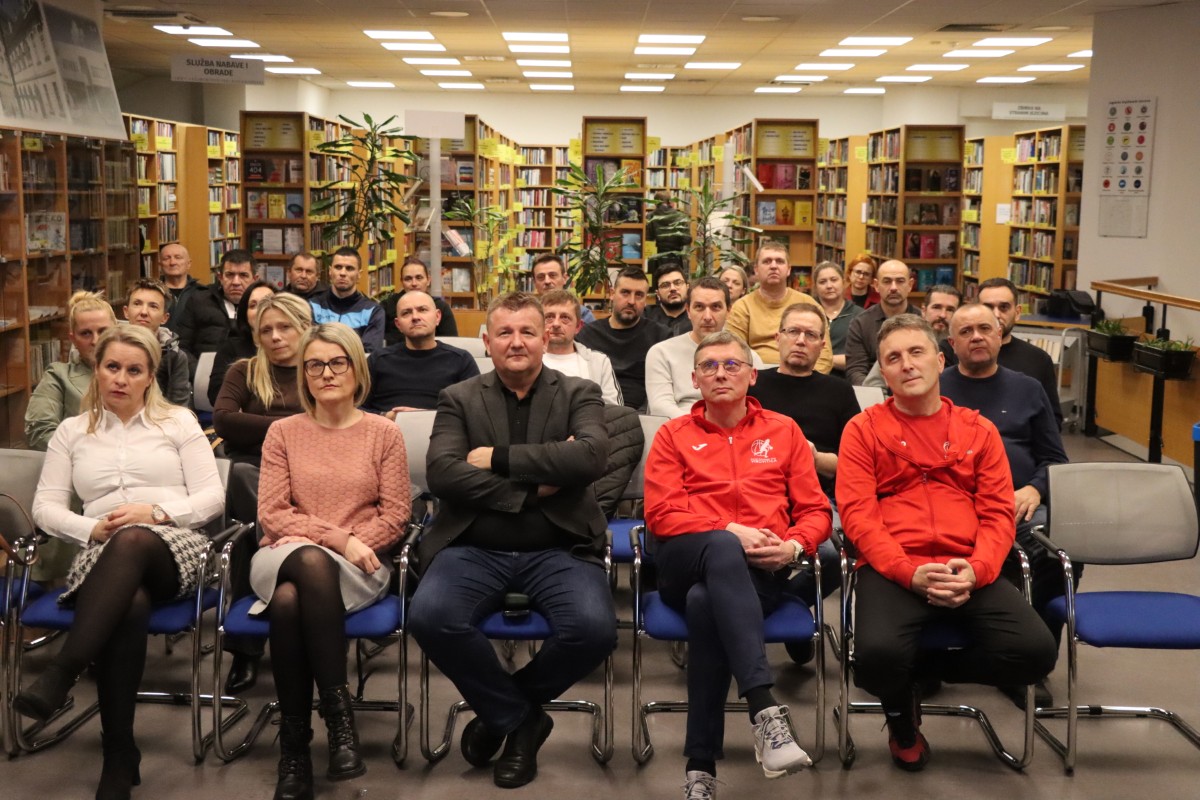 Fotografija prikazuje više ljudi kako sjede u knjižnici i prate neki događaj.