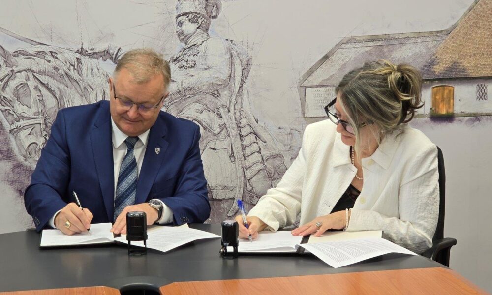 Na fotografiji su dvije osobe koje sjede za stolom i potpisuju dokumente. Muškarac s lijeve strane nosi plavo odijelo i kravatu s prugama, dok žena s desne strane ima bijeli sako i naočale. Na stolu između njih nalaze se dva crna pečata, što dodatno naglašava službeni karakter događaja. U pozadini se vidi mural ili zidna slika s povijesnim motivom: prikazan je lik u tradicionalnoj odjeći na konju, uz građevinu sa slamnatim krovom. Cjelokupna atmosfera fotografije sugerira formalnu prigodu, vjerojatno potpisivanje ugovora, sporazuma ili partnerstva između institucija.