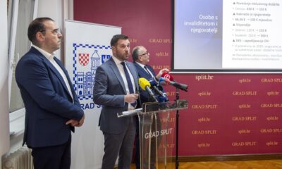 Fotografija prikazuje službenu press‑konferenciju u Splitu, u prostoru gradske uprave. Trojica muškaraca u odijelima stoje iza govornice s nizom mikrofona, što upućuje na javnu objavu ili predstavljanje novih mjera. Iza njih se nalazi službeni pano s grbom i natpisom “GRAD SPLIT”, kao i projekcijsko platno s informacijama o podršci osobama s invaliditetom.