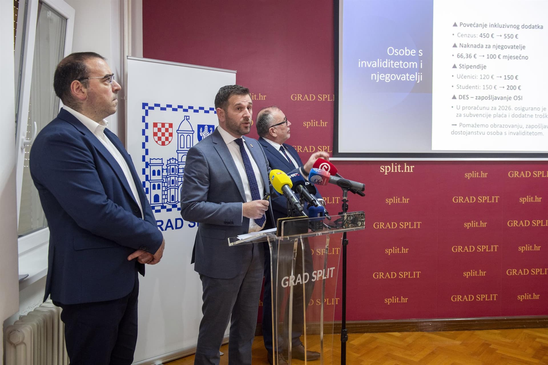 Fotografija prikazuje službenu press‑konferenciju u Splitu, u prostoru gradske uprave. Trojica muškaraca u odijelima stoje iza govornice s nizom mikrofona, što upućuje na javnu objavu ili predstavljanje novih mjera. Iza njih se nalazi službeni pano s grbom i natpisom “GRAD SPLIT”, kao i projekcijsko platno s informacijama o podršci osobama s invaliditetom.