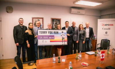 Na fotografiji je devetero ljudi okupljeno u konferencijskoj prostoriji, svi nasmijani i okrenuti prema kameri. U rukama drže veliki simbolični ček namijenjen Institutu Ruđer Bošković u iznosu od 15.000 eura, prikupljen u sklopu humanitarne utrke Terry Fox Run. Iza njih se nalazi zid s umjetničkim slikama i klima‑uređajem, a s desne strane stoji roll‑up pano s logotipima organizatora i partnera, među kojima su Kanada, Esplanade Zagreb Hotel i MPG Inspired Moments. Na stolu ispred grupe vide se boce vode i soka, mali kanadski zastavica te pladanj s grickalicama, što dodatno naglašava svečani, ali opušteni karakter događaja.