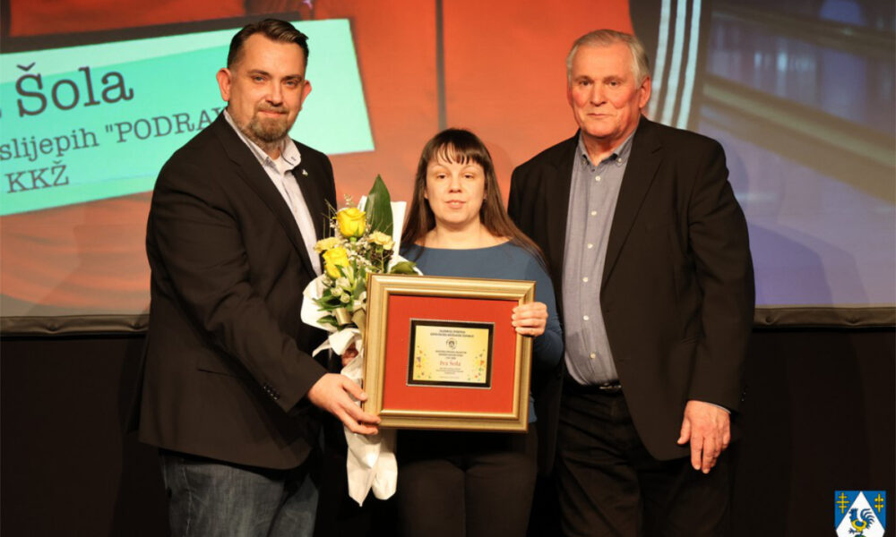 Dodjela priznanja: žena prima nagradu i cvijeće uz dva predstavnika na pozornici.