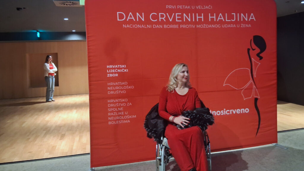 Žena u crvenoj haljini sjedi u invalidskim kolicima ispred službenog panoa Dana crvenih haljina. U pozadini stoji još jedna žena u ležernoj odjeći. Pano sadrži poruke o borbi protiv moždanog udara kod žena te logotipe organizatora.