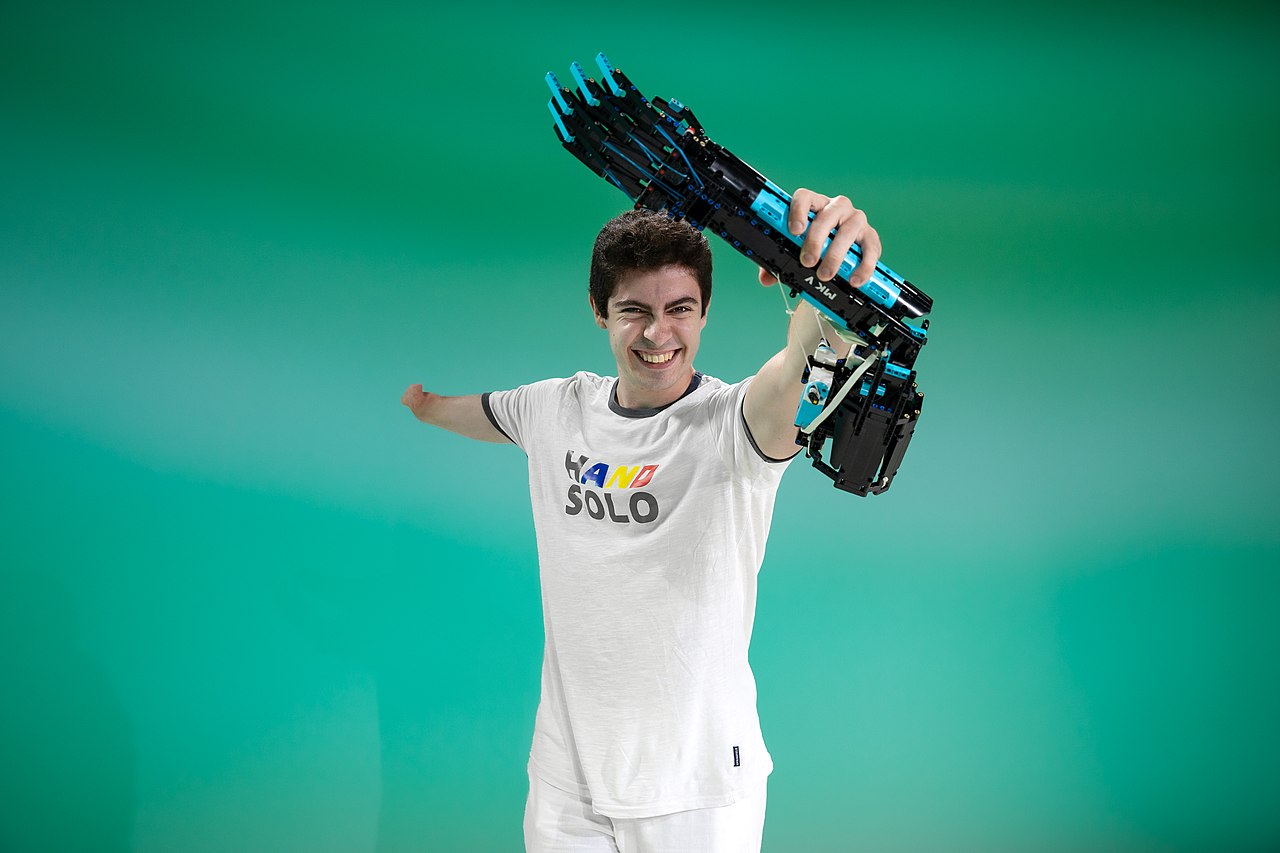 Mlađi muškarac širokog osmijeha pozira ispruženih ruku, pri čemu na desnoj ruci nosi vlastoručno izrađenu robotsku protezu od LEGO kockica. Na bijeloj majici koju nosi piše “HAND SOLO” u šarenim slovima. Pozadina je jednobojna, u zelenim tonovima.