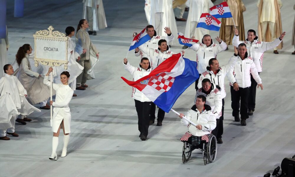 Fotografija prikazuje više ljudi na ceremoniji, a jedan čovjek bez nogu u kolicima nosi hrvatsku zastavu.