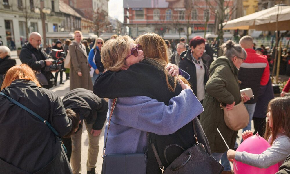Na fotografiji je prikazana živahna gradska ulica puna ljudi. U prvom planu dvije se žene srdačno grle, kao da se dugo nisu vidjele. Jedna nosi svijetloplavi kaput i sunčane naočale, druga tamni kaput i dugu kosu; obje imaju torbice preko ramena. Oko njih se odvija svakodnevna gužva: ljudi prolaze, neki se saginju po stvari, a dijete drži veliki ružičasti balon i štap. U pozadini se vide gradske zgrade, veliki reklamni plakat s licem žene i natkrivena konstrukcija koja podsjeća na trgovačko ili javno područje. Cijela scena djeluje toplo i spontano, naglašavajući trenutak ljudske povezanosti usred užurbanog urbanog okruženja.