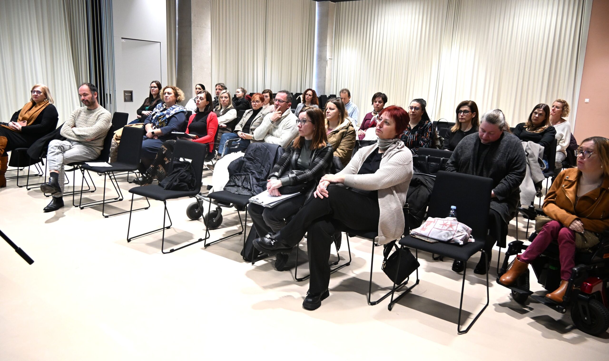 Fotografija prikazuje skupinu odraslih ljudi koji sjede u učionici ili dvorani za predavanja. Svi su okrenuti prema prednjem dijelu prostorije, kao da slušaju predavanje, prezentaciju ili radionicu.