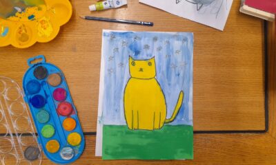 Na fotografiji je prikazan topao, razigran prizor dječje likovne radionice. Na drvenom stolu nalazi se dovršena slika mačke naslikane vodenim bojama. Mačka je žuta, jednostavno i simpatično stilizirana, sjedi na zelenoj travi, a iza nje je plavo nebo ispunjeno ručno nacrtanim zvjezdicama. Oko slike rasuti su razni likovni materijali: paleta s vodenim bojama, žuta posudica s ostacima boje, kistovi, olovka, tube boje, čaša s vodom te papir s početnim skicama mačke. Sve djeluje spontano, kreativno i veselo — kao trenutak u kojem se dijete potpuno uživilo u slikanje.