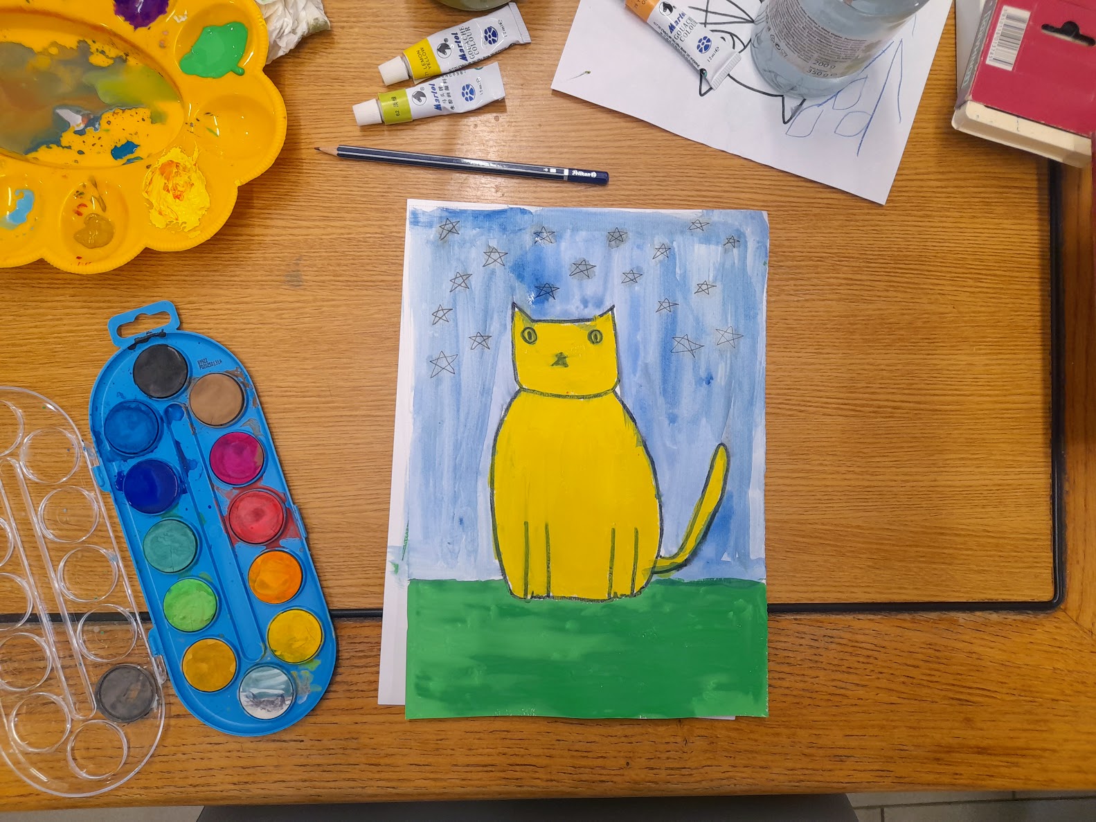 Na fotografiji je prikazan topao, razigran prizor dječje likovne radionice. Na drvenom stolu nalazi se dovršena slika mačke naslikane vodenim bojama. Mačka je žuta, jednostavno i simpatično stilizirana, sjedi na zelenoj travi, a iza nje je plavo nebo ispunjeno ručno nacrtanim zvjezdicama. Oko slike rasuti su razni likovni materijali: paleta s vodenim bojama, žuta posudica s ostacima boje, kistovi, olovka, tube boje, čaša s vodom te papir s početnim skicama mačke. Sve djeluje spontano, kreativno i veselo — kao trenutak u kojem se dijete potpuno uživilo u slikanje.