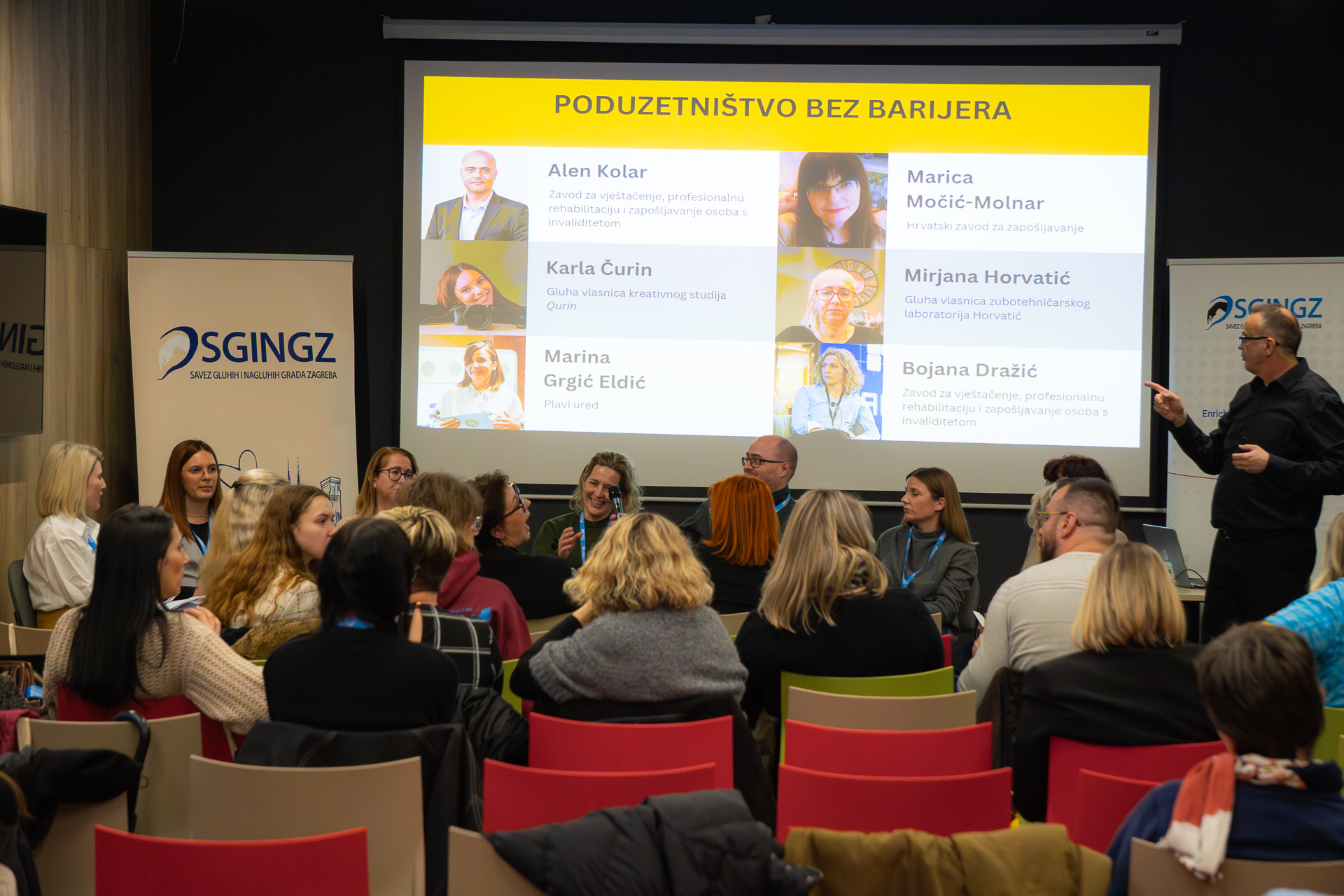 Na fotografiji se vidi dvorana u kojoj se održava panel‑rasprava ili edukativno predavanje. Publika sjedi i prati izlaganje, a s desne strane stoji osoba koja moderira ili predstavlja govornike, pokazujući prema velikom projekcijskom platnu. Na platnu je naslov “Poduzetništvo bez barijera”, uz fotografije i imena šest sudionika te njihove institucije ili poduzeća. Među njima su predstavnici Zavoda za vještačenje, profesionalnu rehabilitaciju i zapošljavanje osoba s invaliditetom, Hrvatskog zavoda za zapošljavanje, Plavog ureda te dvije gluhe poduzetnice koje vode vlastite obrte. Cijela scena djeluje profesionalno i usmjereno na osnaživanje osoba s invaliditetom u području poduzetništva, uz naglasak na pristupačnost i uklanjanje prepreka.