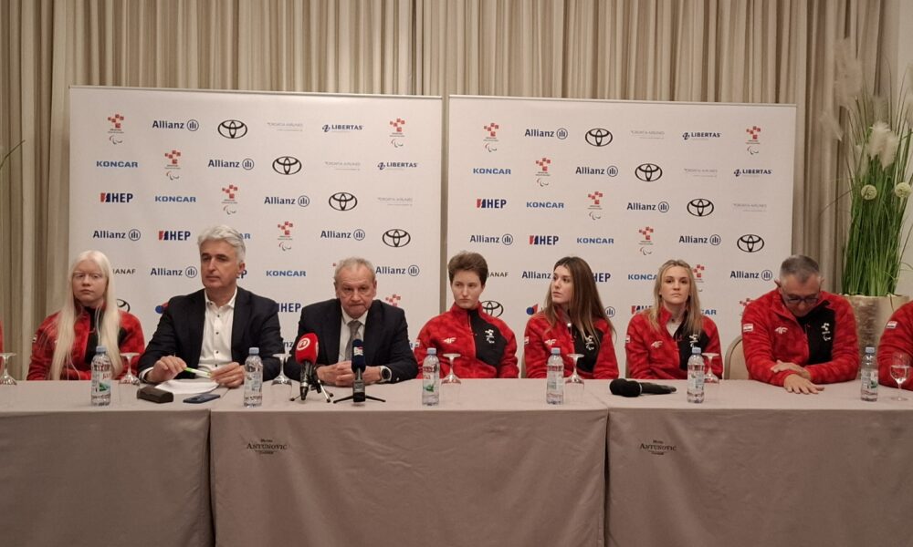 Fotografija prikazuje nekoliko ljudi koji sjede za stolom. Većina njih je u crvenim trenirkama, dok su dvojica ljudi u odjelima.