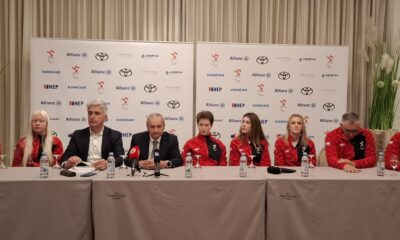 Fotografija prikazuje nekoliko ljudi koji sjede za stolom. Većina njih je u crvenim trenirkama, dok su dvojica ljudi u odjelima.