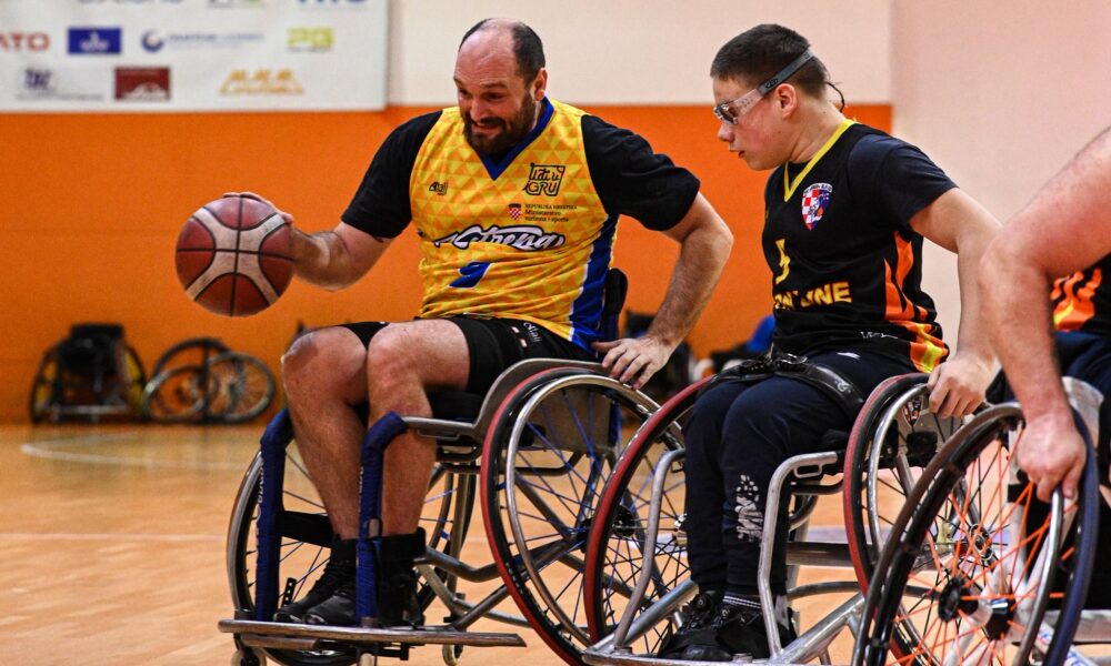 Fotografija šrikazuje utakmicu košarke u kolicima. Jednog igrača kako vodi loptu, dok ga drugi nastoji zaustaviti.