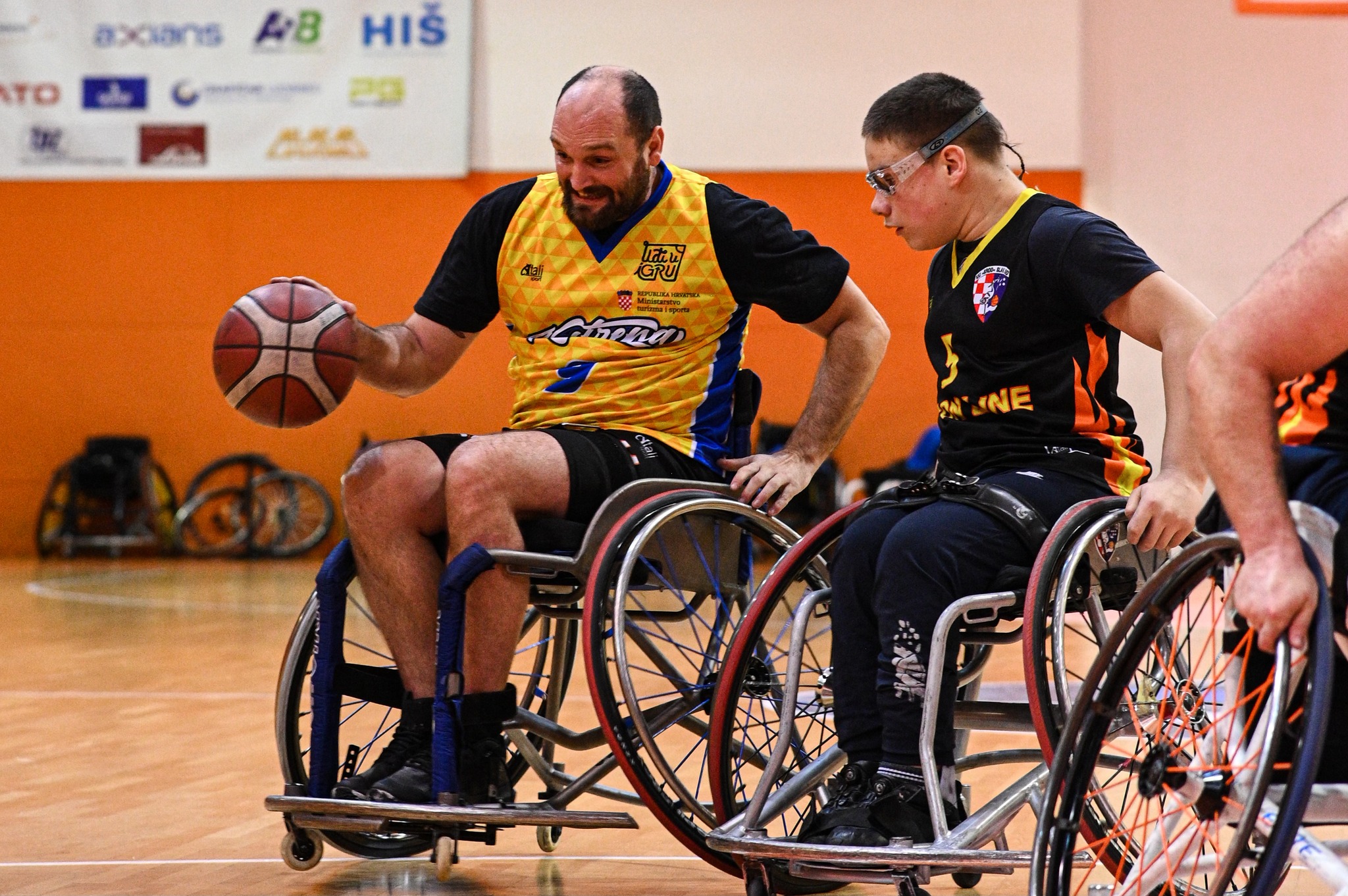 Fotografija šrikazuje utakmicu košarke u kolicima. Jednog igrača kako vodi loptu, dok ga drugi nastoji zaustaviti.