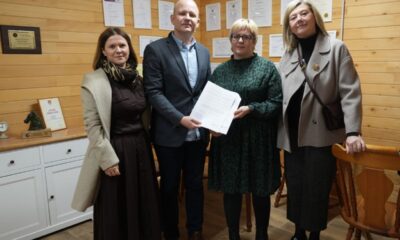 Na fotografiji su četiri odrasle osobe koje stoje u zatvorenom prostoru, u uredu s drvenim zidovima. Dvoje njih – muškarac i žena – zajedno drže dokument, što djeluje kao trenutak potpisivanja, primopredaje ili neke službene potvrde. Svi su odjeveni svečano ili polusvečano, što dodatno naglašava formalnu atmosferu. Iza njih se na zidu nalaze uokvirene diplome, priznanja i nagrade, a s desne strane vidi se bijeli ormarić s još nekoliko uokvirenih dokumenata i malom statuom. Cijeli prostor ostavlja dojam profesionalnog okruženja i važnog događaja koji se upravo obilježava.