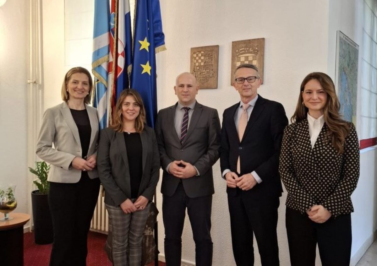 Na fotografiji je pet osoba koje stoje u formalnom, službenom okruženju. Svi su odjeveni u poslovnu odjeću, što stvara dojam službenog sastanka ili diplomatskog događaja. Iza njih se nalaze zastave, među kojima je jasno vidljiva zastava Europske unije, kao i još nekoliko nacionalnih ili regionalnih zastava. Na zidu su postavljeni grbovi, što dodatno naglašava institucionalni ili politički karakter prostora. Cijela scena djeluje kao službena delegacija, potpisivanje sporazuma ili protokolarni susret predstavnika različitih institucija.