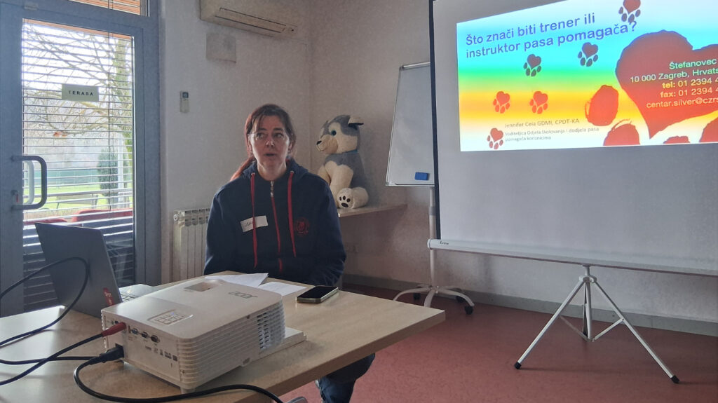 Predavačica sjedi uz projektor i prezentira o ulozi trenera pasa pomagača.