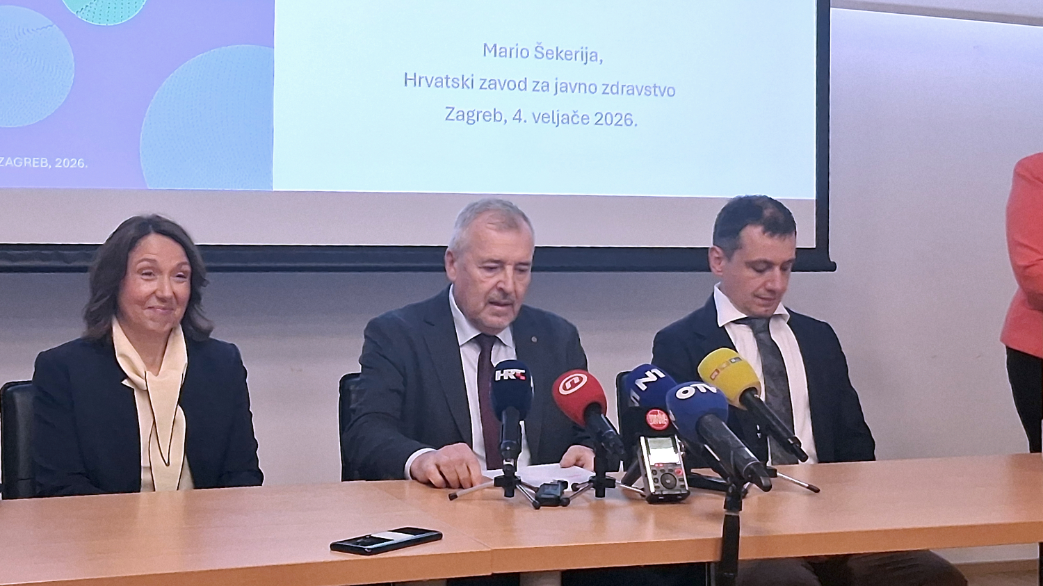 Troje govornika sjedi za stolom tijekom konferencije za medije povodom Svjetskog dana borbe protiv raka, 4. veljače 2026. U sredini govori Mario Šekerija iz Hrvatskog zavoda za javno zdravstvo, dok ispred njih stoje brojni mikrofoni i diktafoni s oznakama medija. U pozadini je prezentacija s naslovom i datumom događaja. Atmosfera je ozbiljna i službena.