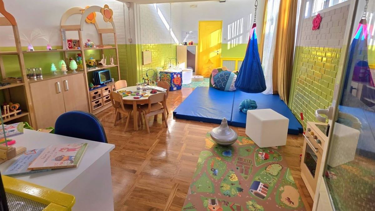 Fotografija prikazuje vedru, moderno uređenu sobu namijenjenu maloj djeci – kombinaciju igraonice i senzorno‑edukativnog prostora. Prostor je svijetao zahvaljujući velikim prozorima, a zidovi su u bijeloj i zelenoj boji, što stvara ugodnu i smirujuću atmosferu.