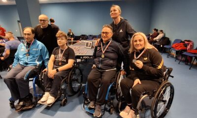 Fotografija prikazuje troje ljude u invalidskim kolicima s medaljama oko vrata. Iznad njih stoji i s njima se slika dvoje ljudi.