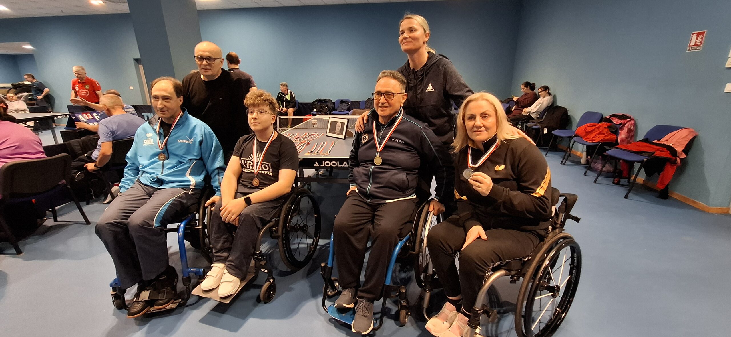 Fotografija prikazuje troje ljude u invalidskim kolicima s medaljama oko vrata. Iznad njih stoji i s njima se slika dvoje ljudi.