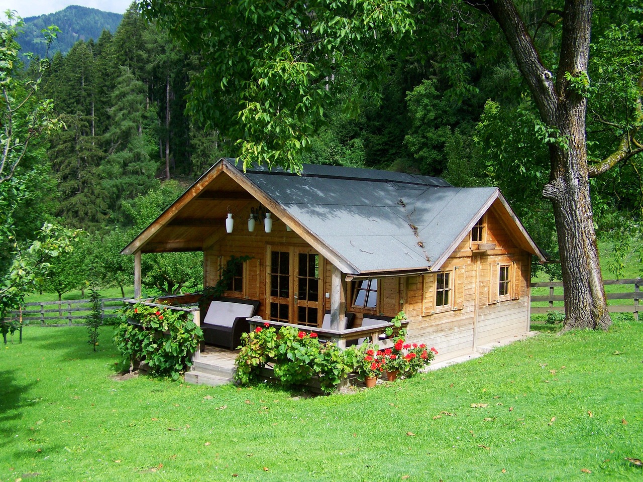 Na slici je prikazana mala drvena kućica smještena usred bujne, zelene prirode. Kućica ima nakošeni krov koji izgleda pomalo istrošeno, a ispred se nalazi mala veranda ukrašena lončanicama i jednostavnim vrtnim namještajem. Oko kućice raste gusta, živopisna trava, a cijeli prostor ograđen je drvenom ogradom. Iza nje se uzdižu visoka stabla i šuma koja prelazi u brdoviti krajolik, što stvara dojam mirnog, skrovitog mjesta idealnog za odmor i bijeg od gradske vreve.