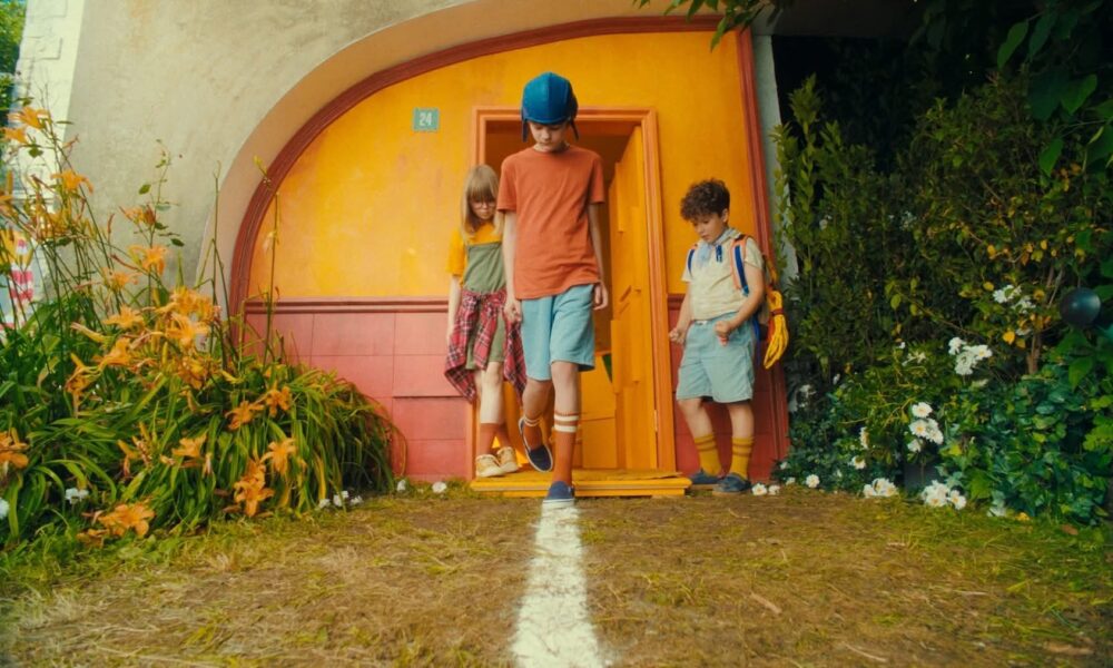 Fotografija prikazuje troje djece kako izlaze iz živopisne kuće jarkih boja. Kuća ima žuta pročelja i narančasta vrata, a iznad njih stoji broj 24. Djeca hodaju prema van po stazi na kojoj je nacrtana bijela linija, što prizoru daje dojam igre ili zamišljene avanture. Središnje dijete nosi plavu kacigu, narančastu majicu, plave kratke hlače i narančaste čarape do koljena. Dijete s lijeve strane obučeno je u žutu majicu, a oko struka ima zavezanu crveno‑kariranu košulju. Dijete s desne strane nosi svijetložutu majicu, plave kratke hlače, žute čarape i na leđima ruksak. Okruženje je puno zelenila i šarenog cvijeća, što cijeloj sceni daje razigran, gotovo bajkovit ugođaj.