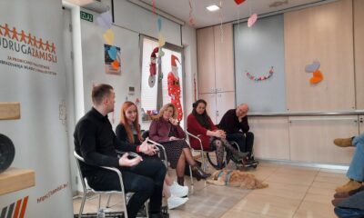 Fotografija prikazuje ugodnu, neformalnu grupnu raspravu u zatvorenom prostoru, ukrašenom visećim šarenim i srcolikim ukrasima. Pet osoba sjedi u polukrugu i razgovara – jedna osoba upravo govori, dok ostali pažljivo slušaju. Ispred njih, na podu, leži pas s oprsnicom, miran i opušten. S lijeve strane stoji roll‑up plakat udruge “Zamisli”, koja se bavi promicanjem kvalitetnog obrazovanja mladih s invaliditetom. U pozadini se vidi i izlazna oznaka “Izlaz”. Cijela scena djeluje inkluzivno, prijateljski i usmjereno na zajednički rad ili razmjenu iskustava.