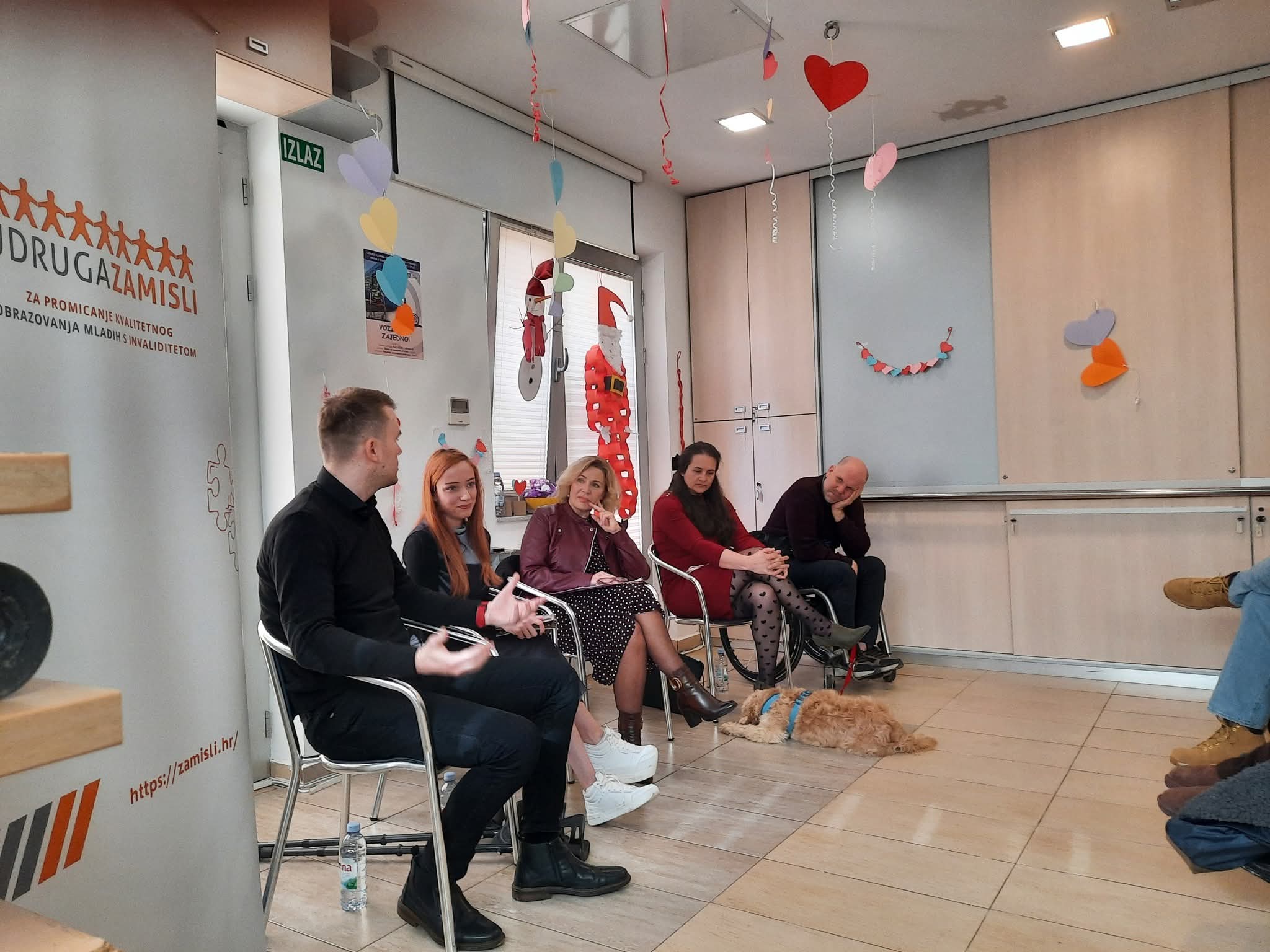 Fotografija prikazuje ugodnu, neformalnu grupnu raspravu u zatvorenom prostoru, ukrašenom visećim šarenim i srcolikim ukrasima. Pet osoba sjedi u polukrugu i razgovara – jedna osoba upravo govori, dok ostali pažljivo slušaju. Ispred njih, na podu, leži pas s oprsnicom, miran i opušten. S lijeve strane stoji roll‑up plakat udruge “Zamisli”, koja se bavi promicanjem kvalitetnog obrazovanja mladih s invaliditetom. U pozadini se vidi i izlazna oznaka “Izlaz”. Cijela scena djeluje inkluzivno, prijateljski i usmjereno na zajednički rad ili razmjenu iskustava.