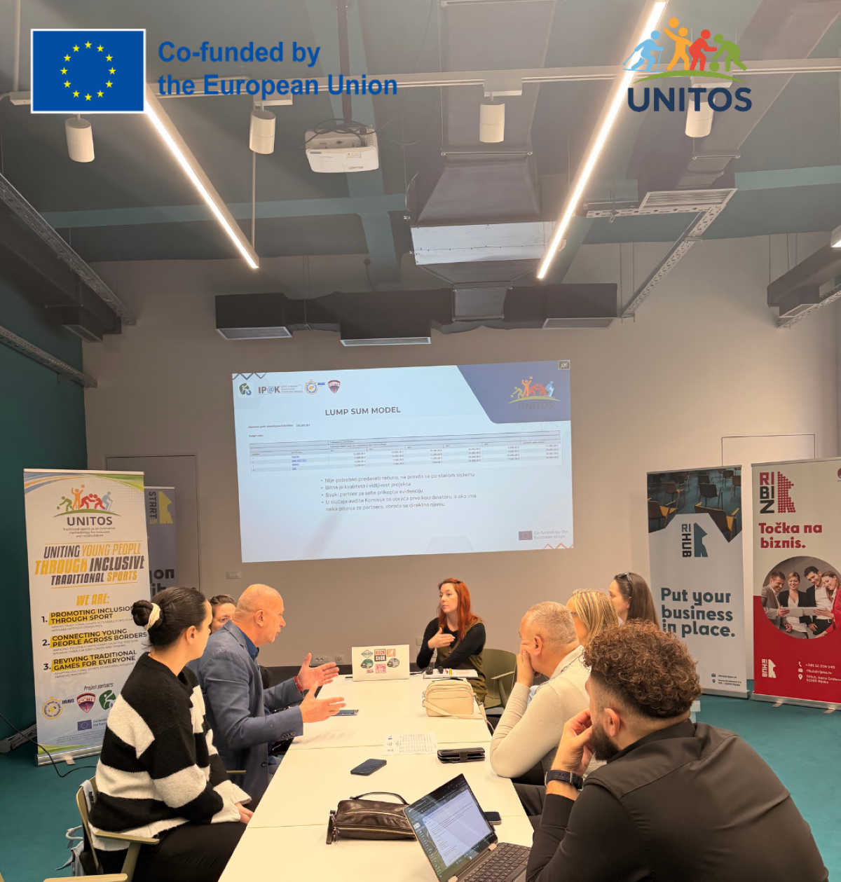 Na fotografiji je šest osoba koje sjede oko konferencijskog stola u modernoj, dobro osvijetljenoj dvorani za sastanke. Atmosfera djeluje profesionalno i usmjerena na zajednički rad, kao da sudionici sudjeluju u radionici, edukaciji ili projektnoj raspravi.