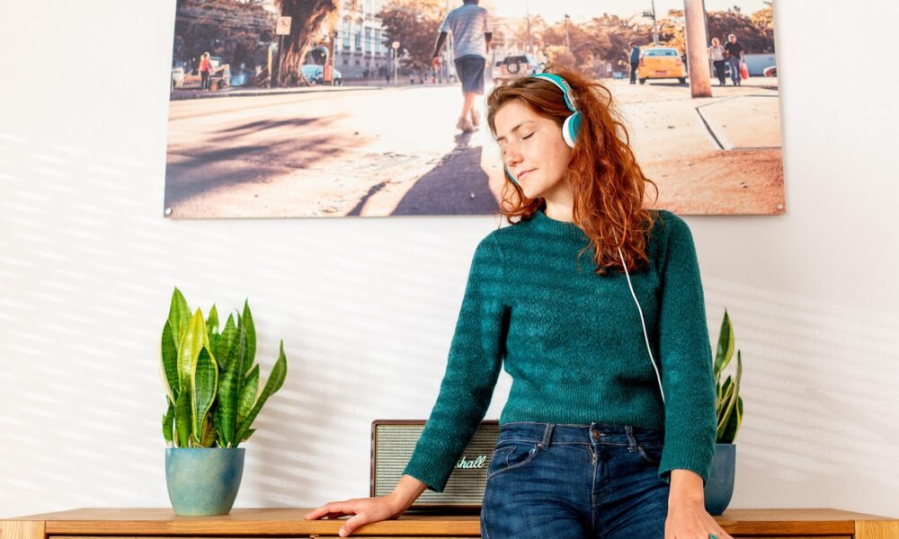 Na slici je osoba s dugom, kovrčavom crvenom kosom koja stoji ispred drvenog ormarića. Nosi plavo‑bijele slušalice i izgleda kao da uživa u glazbi – oči su zatvorene, a izraz lica opušten. Odjevena je u tirkiznu vestu i traperice. Na ormariću se nalaze dvije sanseverije u posudama, postavljene simetrično s obje strane crnog Marshall zvučnika. Iza osobe visi velika fotografija užurbane gradske ulice po sunčanom danu, s ljudima, drvećem, zgradama i žutim taksijem. Cijela scena djeluje topla, ugodna i skladno uređena.