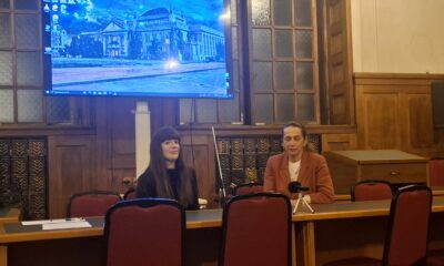 Fotografija prikazuje dvije žene kako sjede za stolom dok se u pozadini vidi kompjutorska pozadina.