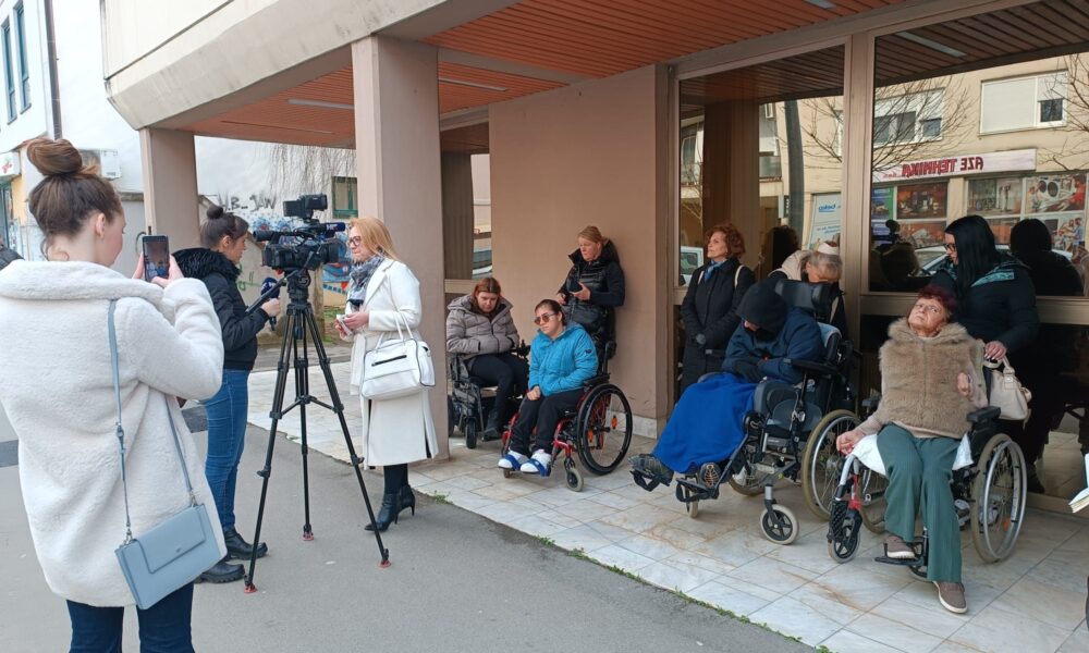 Na fotografiji je prikazan skup ljudi ispred zgrade, vjerojatno tijekom davanja izjave za medije ili konferencije za novinare. U prvom planu vidi se nekoliko novinara i snimatelja s kamerom i mikrofonom, dok jedna žena govori pred kamerom. U pozadini, uz ulaz u zgradu, nalazi se više osoba u invalidskim kolicima, zajedno s pratnjom i drugim okupljenima. Neki od njih promatraju situaciju, dok drugi čekaju ili razgovaraju. Atmosfera djeluje ozbiljno i organizirano, što upućuje na javno obraćanje ili istup vezan uz određeni društveni problem. Cijela scena sugerira da se radi o događaju vezanom uz osobe s invaliditetom i njihove asistente, uz prisutnost medija koji prate situaciju.