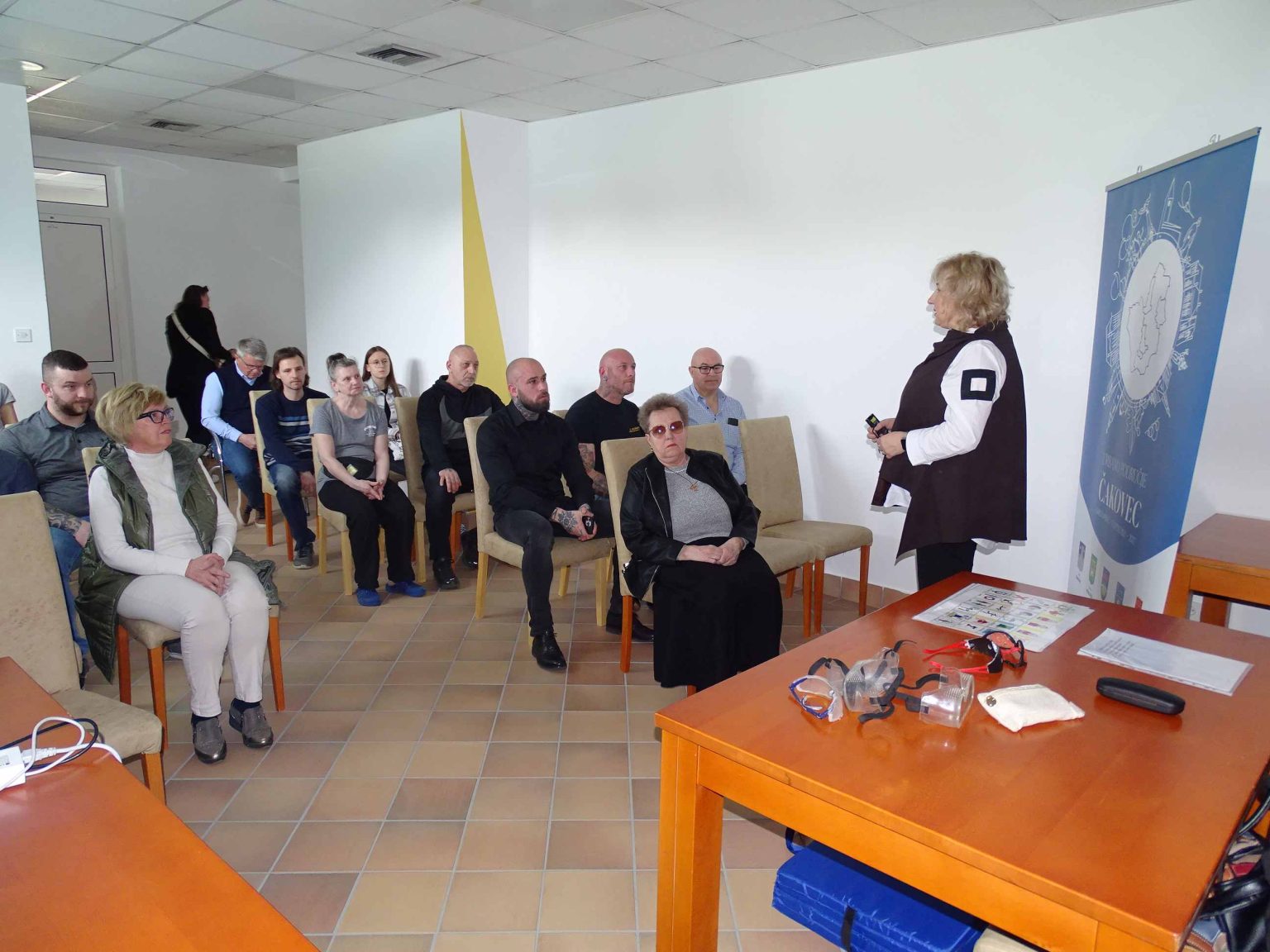 Na fotografiji je prikazana edukacija koja se održava u zatvorenom prostoru, vjerojatno u učionici ili konferencijskoj dvorani. U prvom planu nalazi se žena koja stoji i obraća se okupljenim sudionicima, držeći prezentacijski uređaj u ruci. Uz nju je postavljen roll-up plakat s vizualnim identitetom Grada Čakovca. Sudionici edukacije sjede na stolicama raspoređenima u redove i pažljivo slušaju predavačicu. Riječ je o manjoj skupini odraslih osoba različite dobi. Na stolu ispred predavačice nalaze se razni predmeti, uključujući zaštitne maske i opremu koja se vjerojatno koristi kao dio praktičnog dijela edukacije. Prostorija je jednostavno uređena, svijetlih zidova i s dovoljno prirodnog svjetla, što doprinosi formalnoj, ali ugodnoj atmosferi. Cjelokupan prizor ostavlja dojam organizirane i interaktivne edukacije usmjerene na praktično učenje.