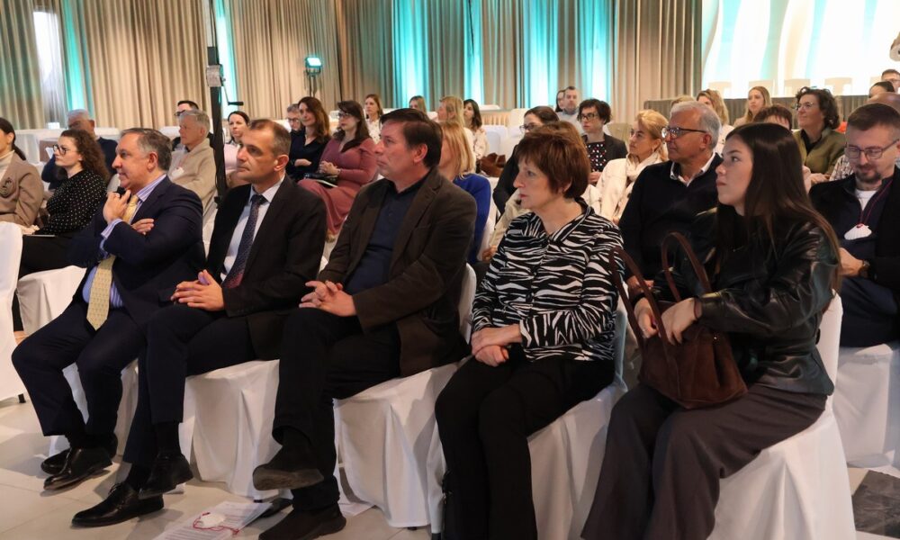 Na fotografiji je prikazan skup ljudi koji sudjeluju na konferenciji ili javnom događanju u zatvorenom prostoru. Sudionici sjede u redovima na bijelim stolcima, okrenuti prema govorniku koji nije vidljiv na slici. U prvom redu nalaze se formalno odjeveni muškarci i žene, što upućuje na službeni ili stručni karakter događaja. Većina prisutnih pažljivo sluša, s ozbiljnim i koncentriranim izrazima lica. Prostorija je moderno uređena, s dugim zavjesama u neutralnim tonovima i ambijentalnim osvjetljenjem koje daje ugodnu atmosferu. U pozadini se vidi veći broj sudionika, što sugerira dobru posjećenost događaja. Cjelokupni dojam fotografije je profesionalan i organiziran, a atmosfera mirna i usmjerena na razmjenu znanja i informacija.