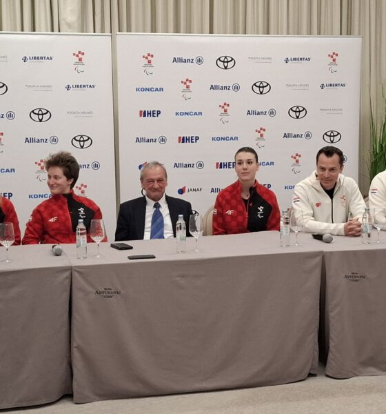 Fotografija prikazuje nekoliko ljudi u crvenim trenirkama, dok je jedan čovjek u odjelu. Iza njih je bilbord.