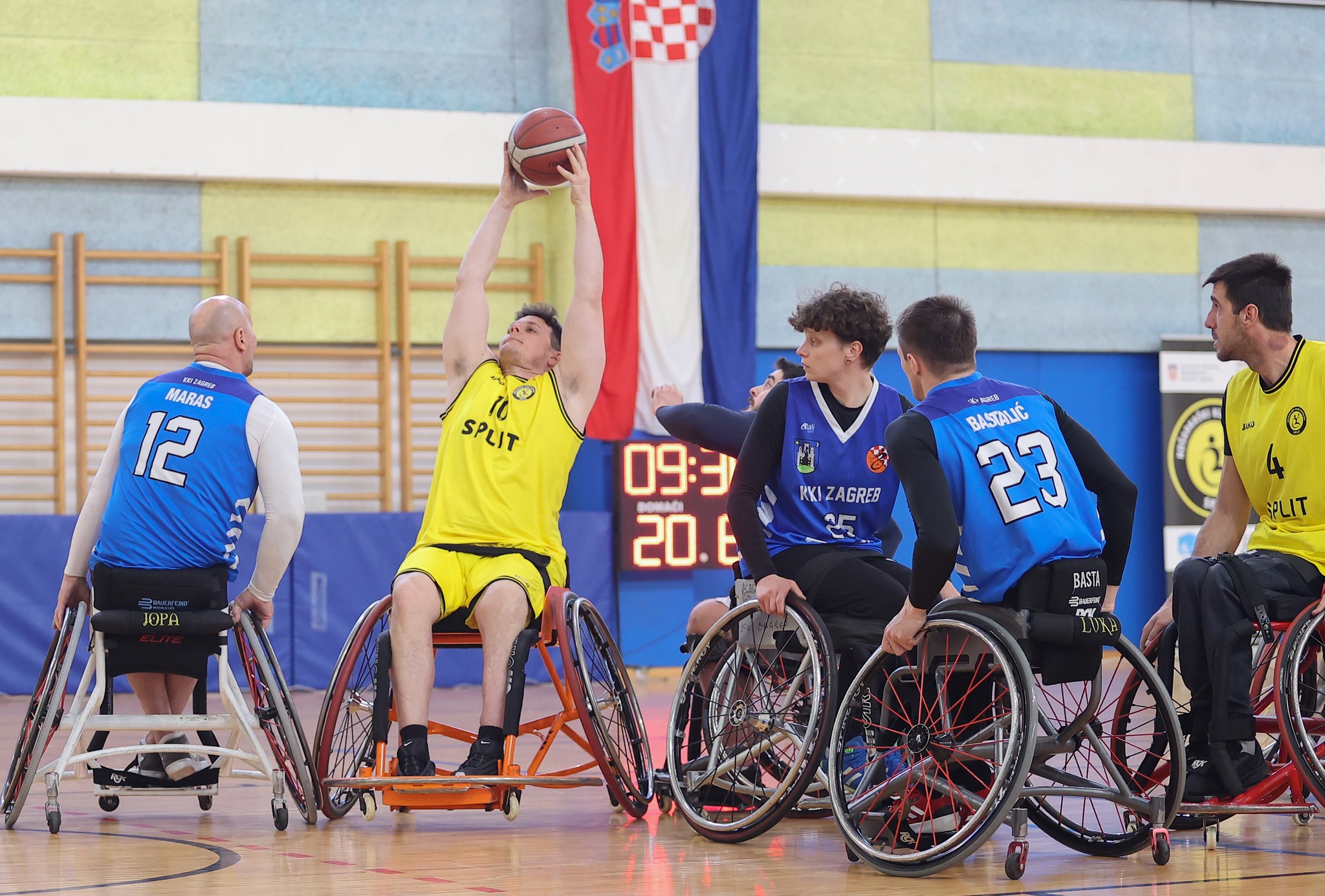 Fotografija prikazuje jedan tim u žutim drugi u plavim dresovima kako igraju košarku u kolicima. Igrač u žutom dresu pokušava baciti loptu, a oko njega je više igrača u plavim dresovima. Na fotografiji se vidi i velika hrvatska zastava.