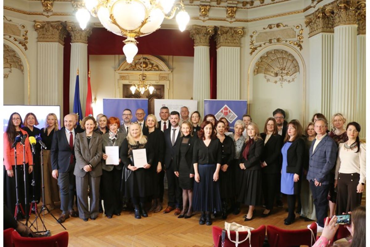 Na slici se nalazi veća skupina ljudi okupljena u svečanoj dvorani bogato ukrašenoj klasičnim elementima poput stupova, ukrasa i velikog lustera. Prostor izgleda reprezentativno, poput kazališta ili važne državne institucije. Ljudi su odjeveni formalno, u poslovnu odjeću, što upućuje na službeni događaj. U prvom redu nekoliko osoba drži dokumente u rukama, vjerojatno ugovore ili potvrde. S lijeve strane vide se mikrofoni, što sugerira da je riječ o javnom događaju ili konferenciji za medije. U pozadini su postavljene zastave i promotivni paneli, a cijela scena djeluje kao svečana dodjela ili obilježavanje važnog događaja, uz prisutnost većeg broja sudionika i predstavnika institucija.