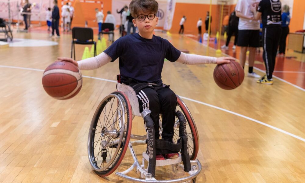 Dječak u kolicima dribla dvije košarkaške lopte u sportskoj dvorani.