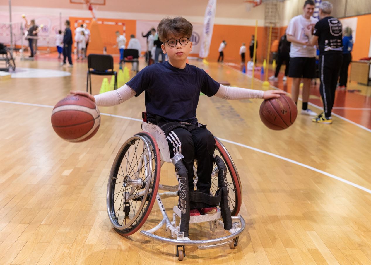Dječak u kolicima dribla dvije košarkaške lopte u sportskoj dvorani.