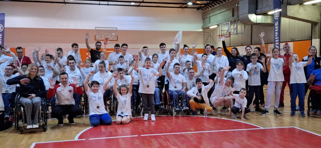 Velika grupa djece i mladih s invaliditetom pozira i maše u dvorani.