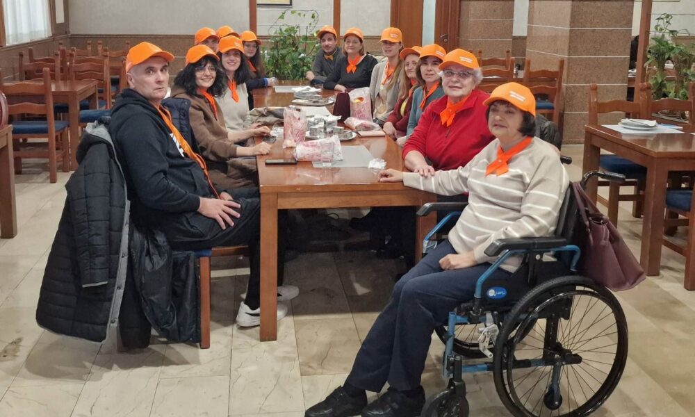 Na fotografiji se vidi veća skupina ljudi okupljena oko drvenog stola u ugodnom, zatvorenom prostoru koji podsjeća na blagovaonicu ili zajedničku prostoriju. Svi nose narančaste kape i marame, što daje dojam da sudjeluju u istom programu, radionici ili timu. Atmosfera djeluje prijateljski i opušteno.