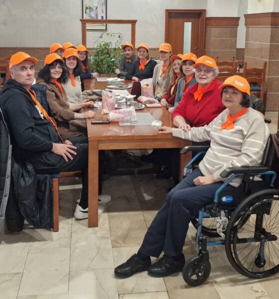 Na fotografiji se vidi veća skupina ljudi okupljena oko drvenog stola u ugodnom, zatvorenom prostoru koji podsjeća na blagovaonicu ili zajedničku prostoriju. Svi nose narančaste kape i marame, što daje dojam da sudjeluju u istom programu, radionici ili timu. Atmosfera djeluje prijateljski i opušteno.