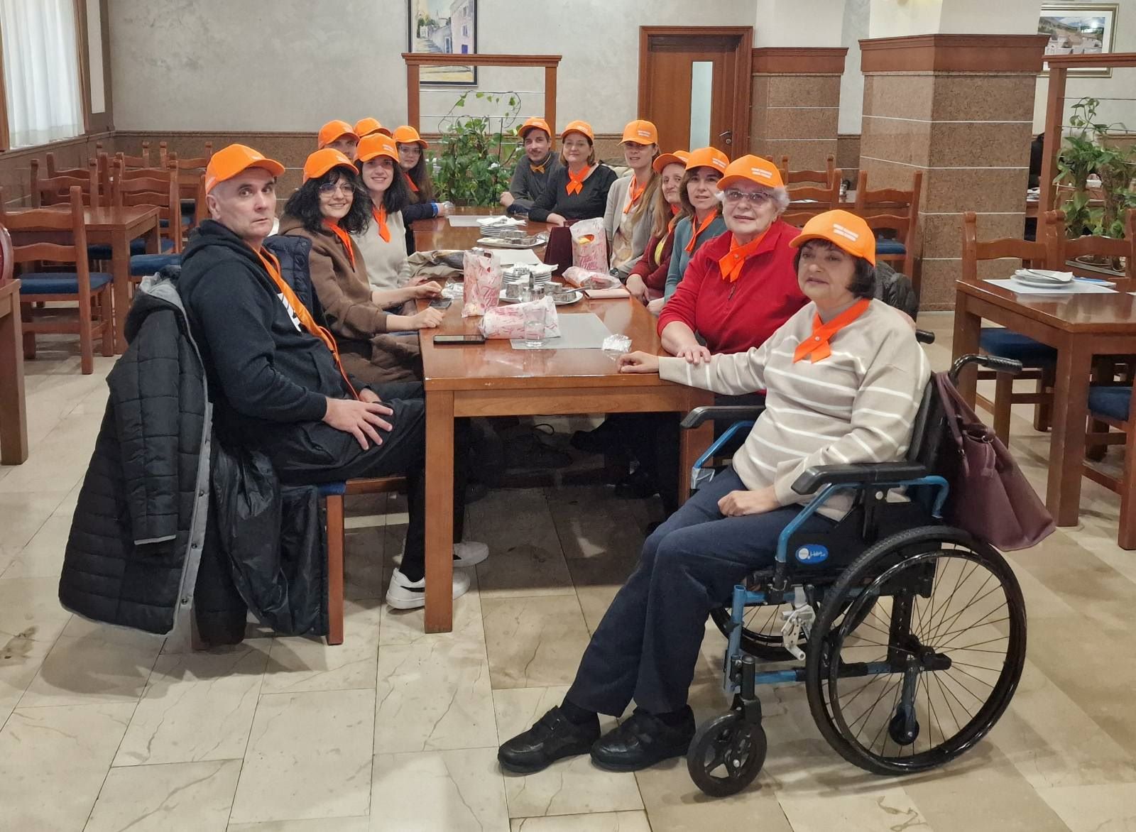 Na fotografiji se vidi veća skupina ljudi okupljena oko drvenog stola u ugodnom, zatvorenom prostoru koji podsjeća na blagovaonicu ili zajedničku prostoriju. Svi nose narančaste kape i marame, što daje dojam da sudjeluju u istom programu, radionici ili timu. Atmosfera djeluje prijateljski i opušteno.